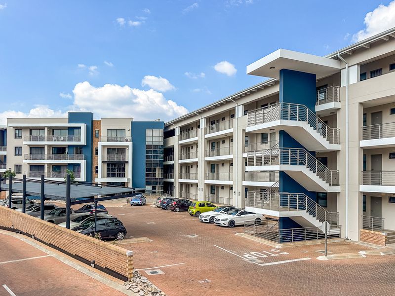 Exterior view of a modern apartment complex featuring multi-level balconies, external staircases, and a paved parking area with covered carports.
