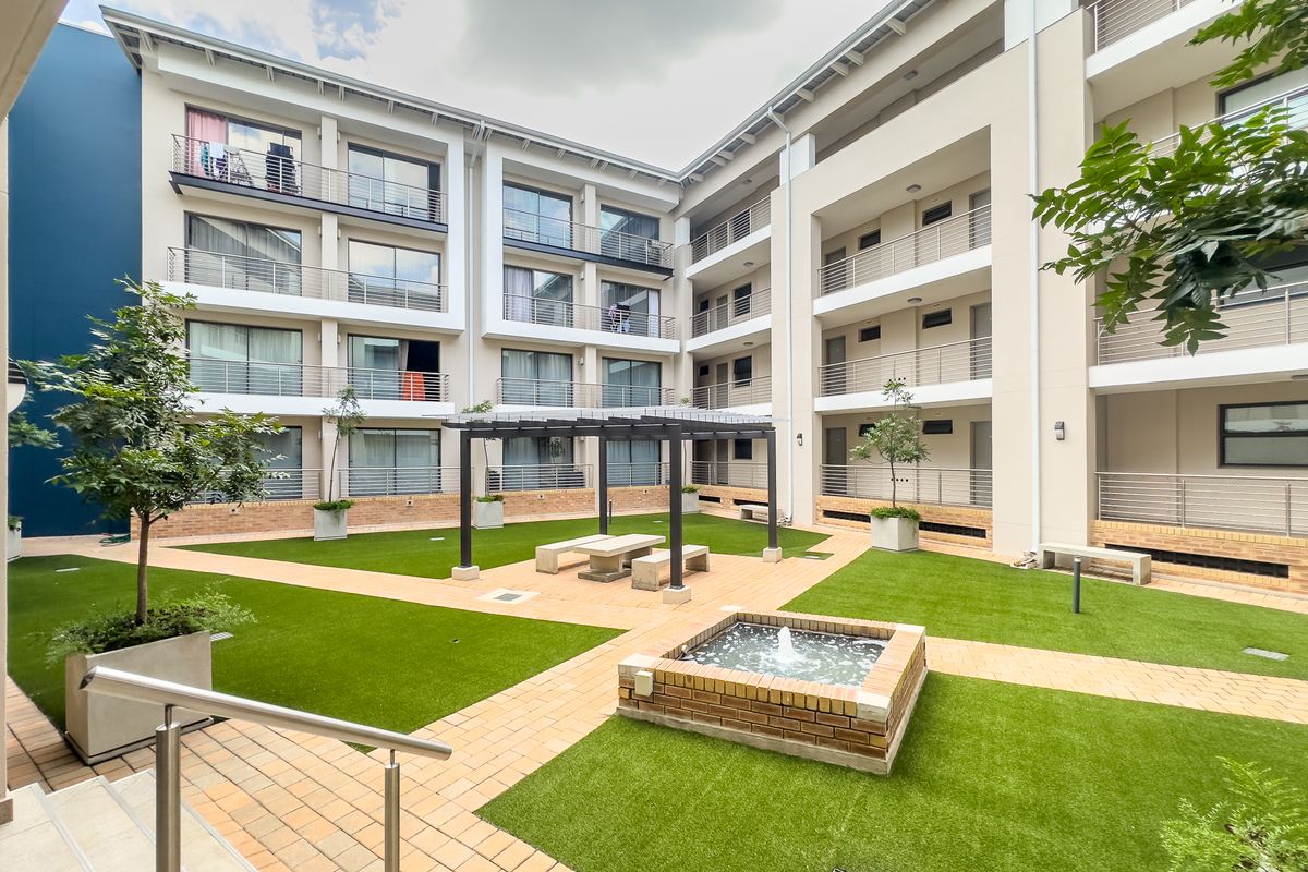 A well-maintained communal courtyard featuring artificial lawn, brick paving, a central water feature, and a pergola, surrounded by multi-story apartment balconies.
