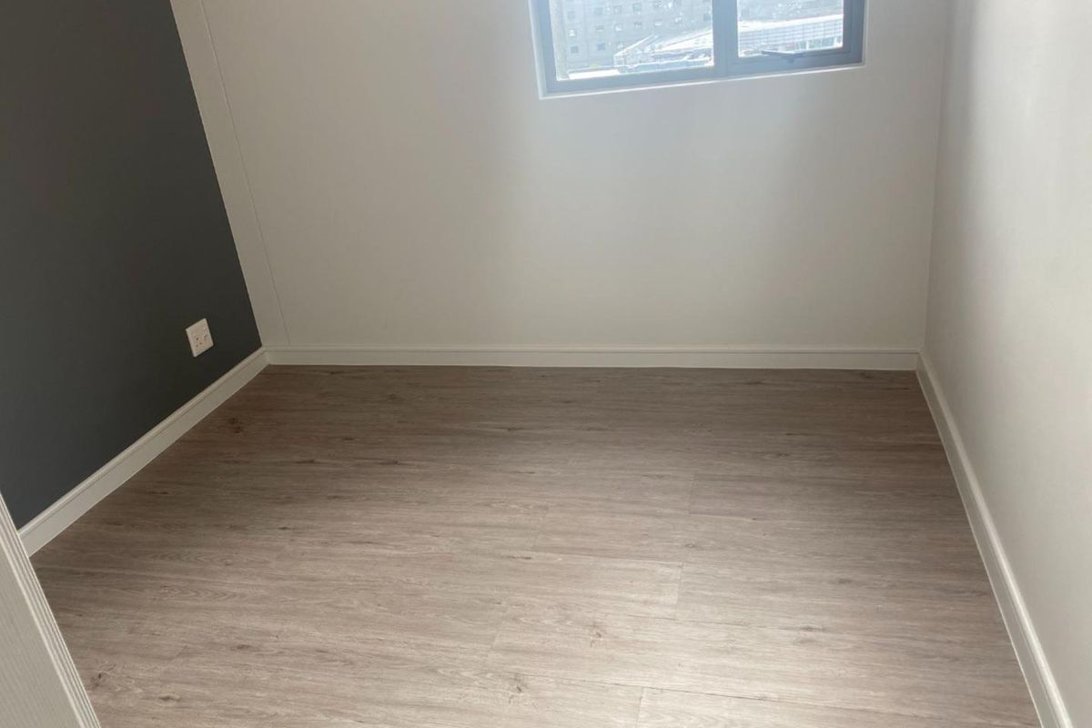 A clean, empty bedroom featuring modern light wood-look laminate flooring, a neutral color palette with a dark accent wall, and a window.