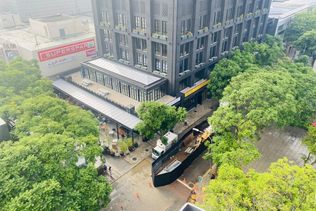Aerial view of a modern multi-story apartment building with ground-level retail, surrounding trees, and pedestrian street access in an urban setting.