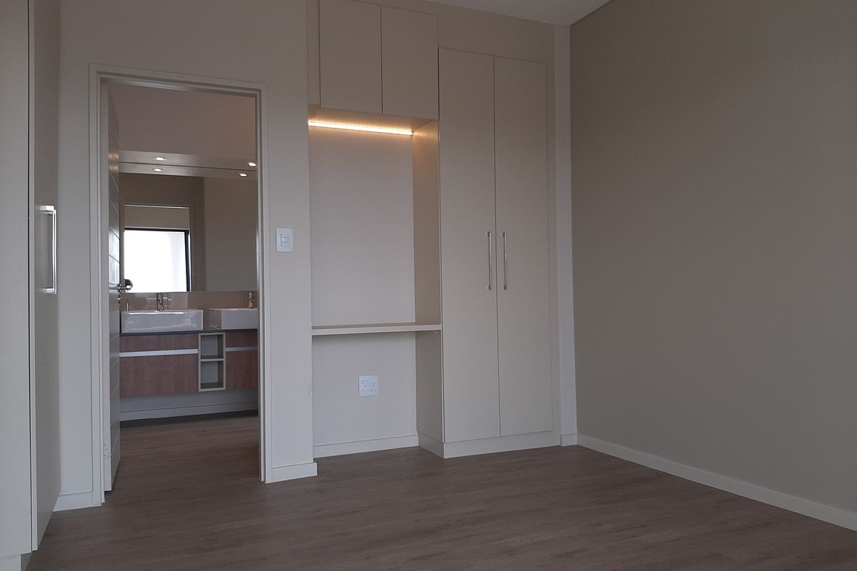 A modern bedroom featuring built-in cabinetry with integrated lighting and a desk area, leading into an en-suite bathroom with double vanity.