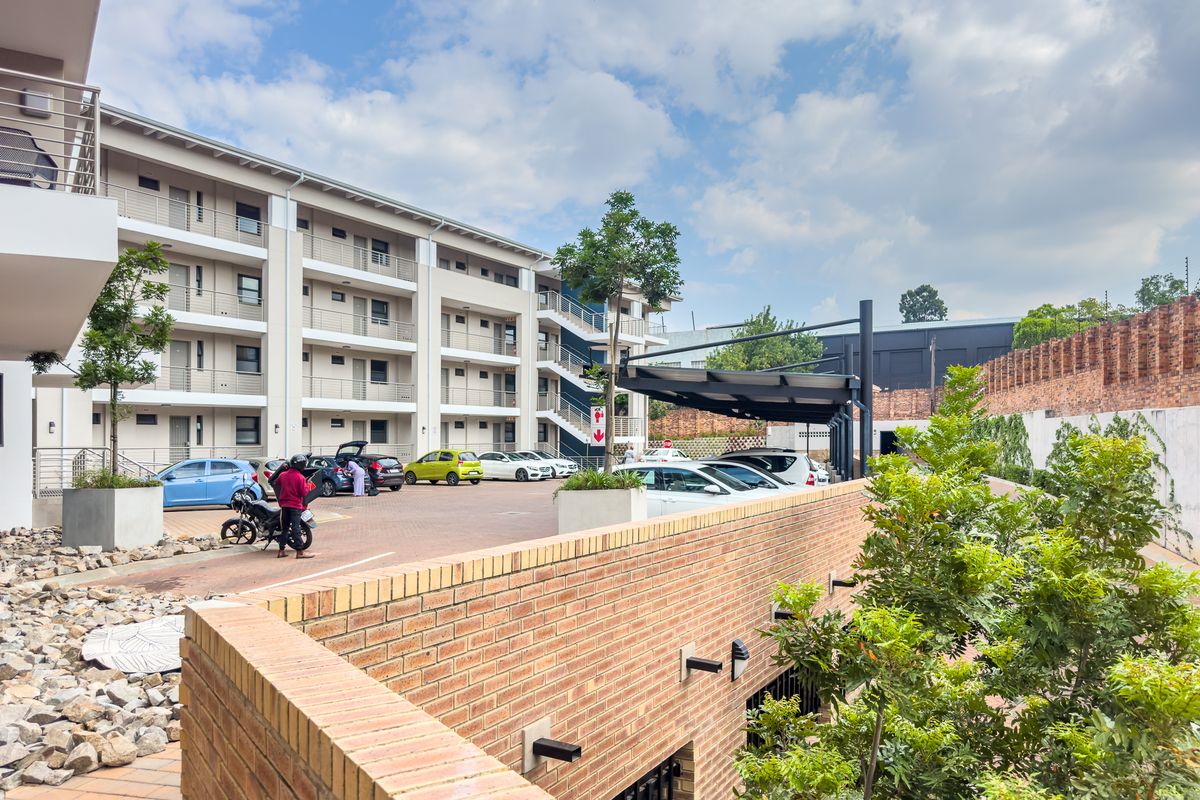 Exterior view of the apartment complex featuring paved parking areas, covered parking bays, and landscaped garden beds with brick retaining walls.