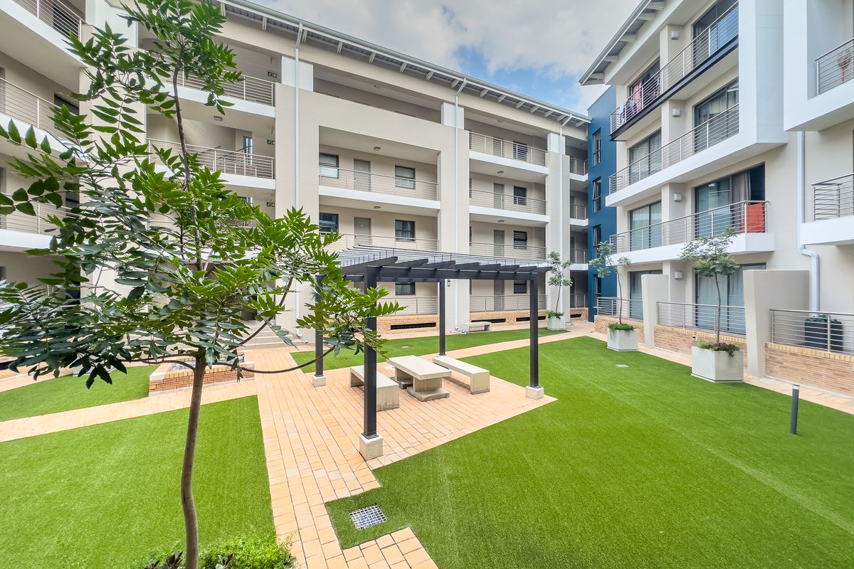 A well-maintained communal courtyard area featuring paved walkways, artificial lawn, and a central pergola with built-in seating, surrounded by modern apartment building facades.
