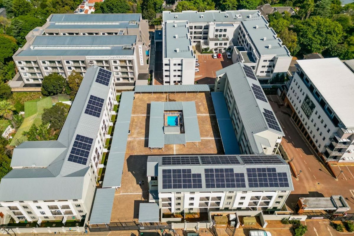 Aerial view of the apartment complex featuring multiple residential buildings, a central swimming pool area, and extensive rooftop solar panel installations.