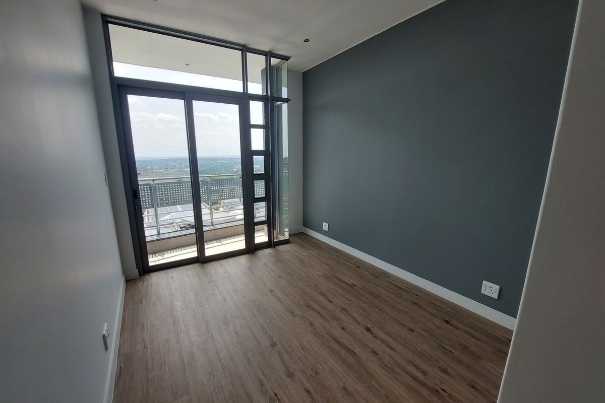 A bright, modern, and empty bedroom featuring laminate flooring, a dark accent wall, and large sliding glass doors leading to a private balcony.