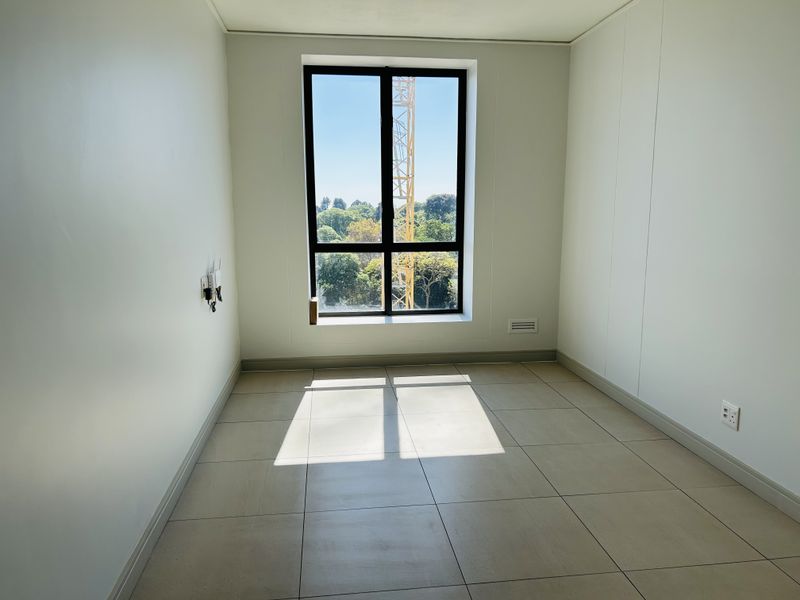 A bright, empty bedroom featuring neutral tiled flooring, clean white walls, and a large window providing natural light and an exterior view.