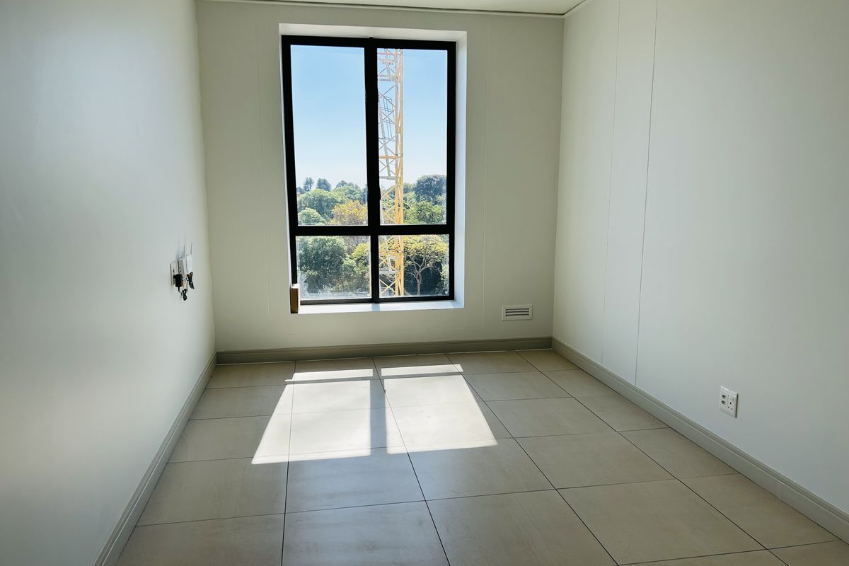 A bright, empty bedroom featuring neutral tiled flooring, clean white walls, and a large window providing natural light and an exterior view.