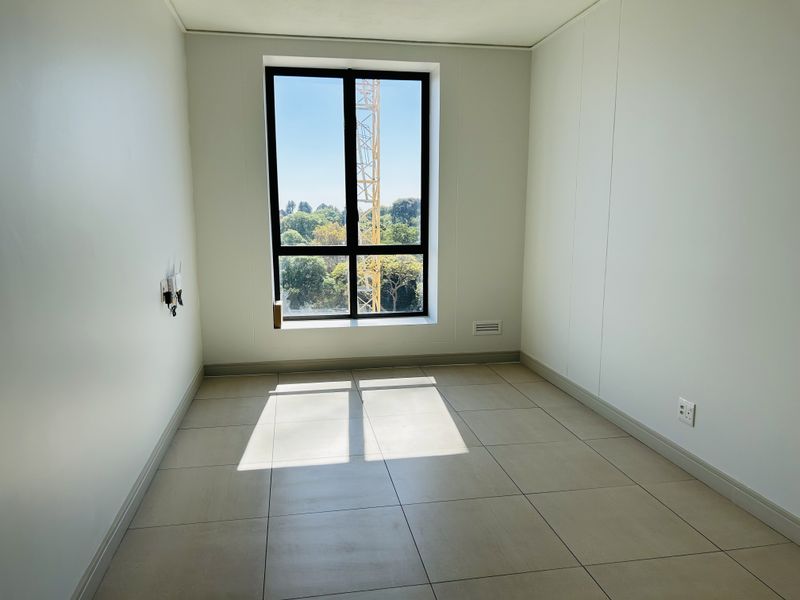 A bright, empty bedroom featuring neutral tiled flooring, clean white walls, and a large window providing natural light and an exterior view.