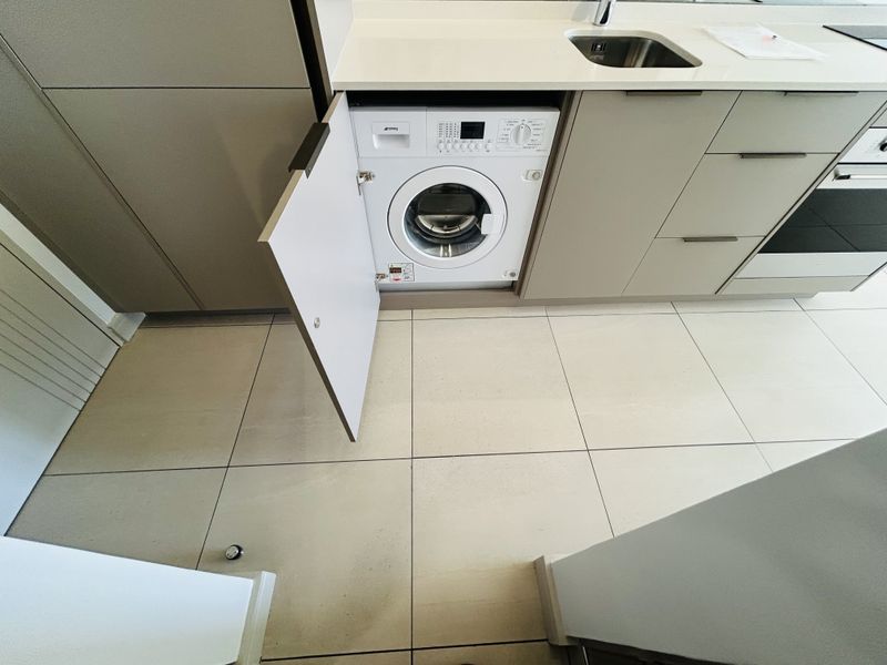 Modern kitchen featuring integrated washing machine, sleek cabinetry, stone countertops, and stainless steel sink with neutral ceramic floor tiling.