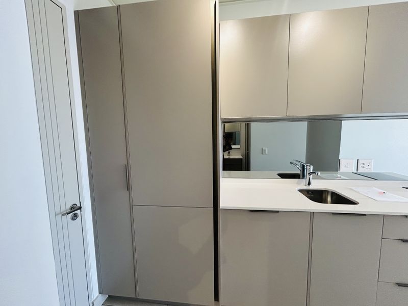 Modern kitchen area featuring sleek taupe cabinetry, stone countertops, an undermount sink with a mixer tap, and a reflective mirrored splashback.