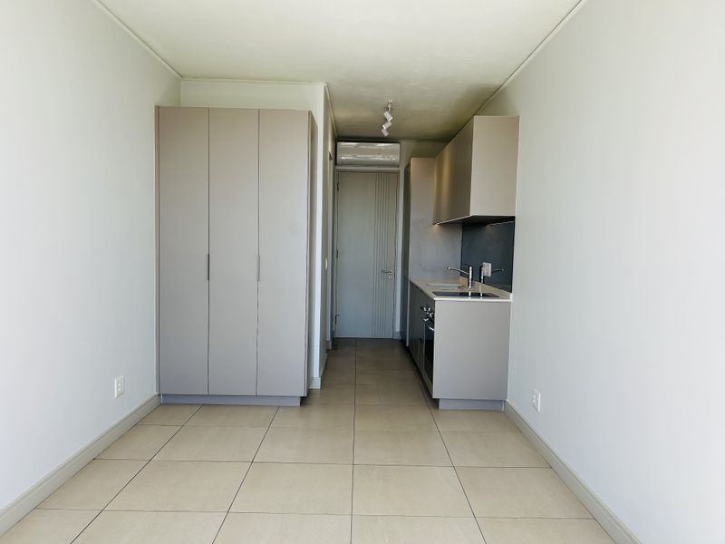 An open-plan studio space featuring a compact kitchenette with built-in cabinetry, integrated appliances, and neutral tiled flooring throughout.