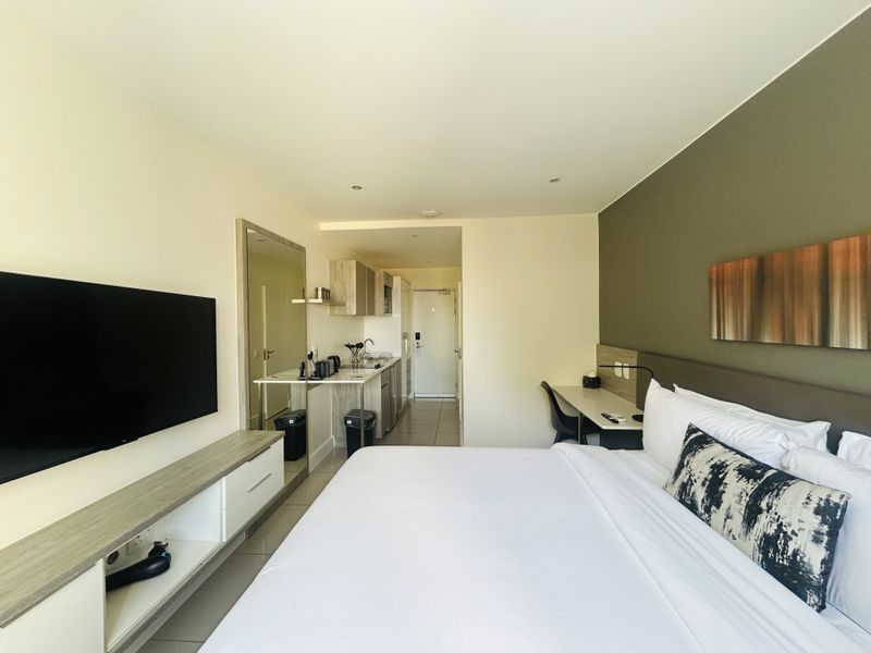 A modern studio bedroom featuring a compact kitchenette, built-in desk, and media console with neutral finishes and recessed lighting.