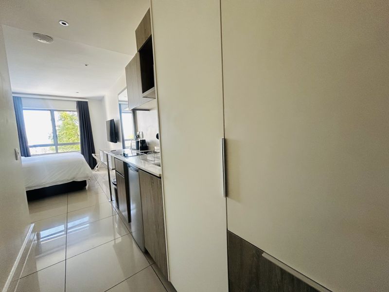 Modern open-plan studio apartment featuring a compact kitchenette with built-in cabinetry, sleek tiled flooring, and a sliding glass door leading to a bright bedroom area.