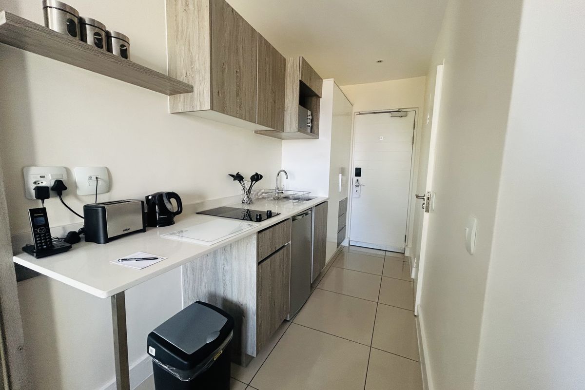 Compact modern kitchen featuring light wood-grain cabinetry, integrated electric hob, stainless steel sink, and neutral tiled flooring, leading directly to the apartment entrance.