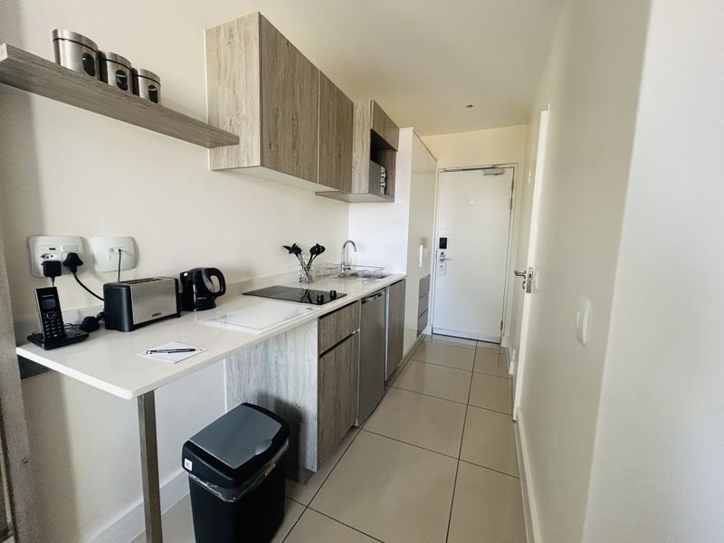 Compact modern kitchen featuring light wood-grain cabinetry, integrated electric hob, stainless steel sink, and neutral tiled flooring, leading directly to the apartment entrance.