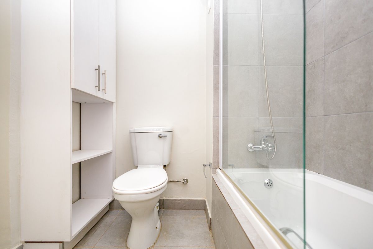 A clean, functional bathroom featuring a bathtub with a glass screen, a toilet, and built-in storage cabinetry with neutral grey tiling.