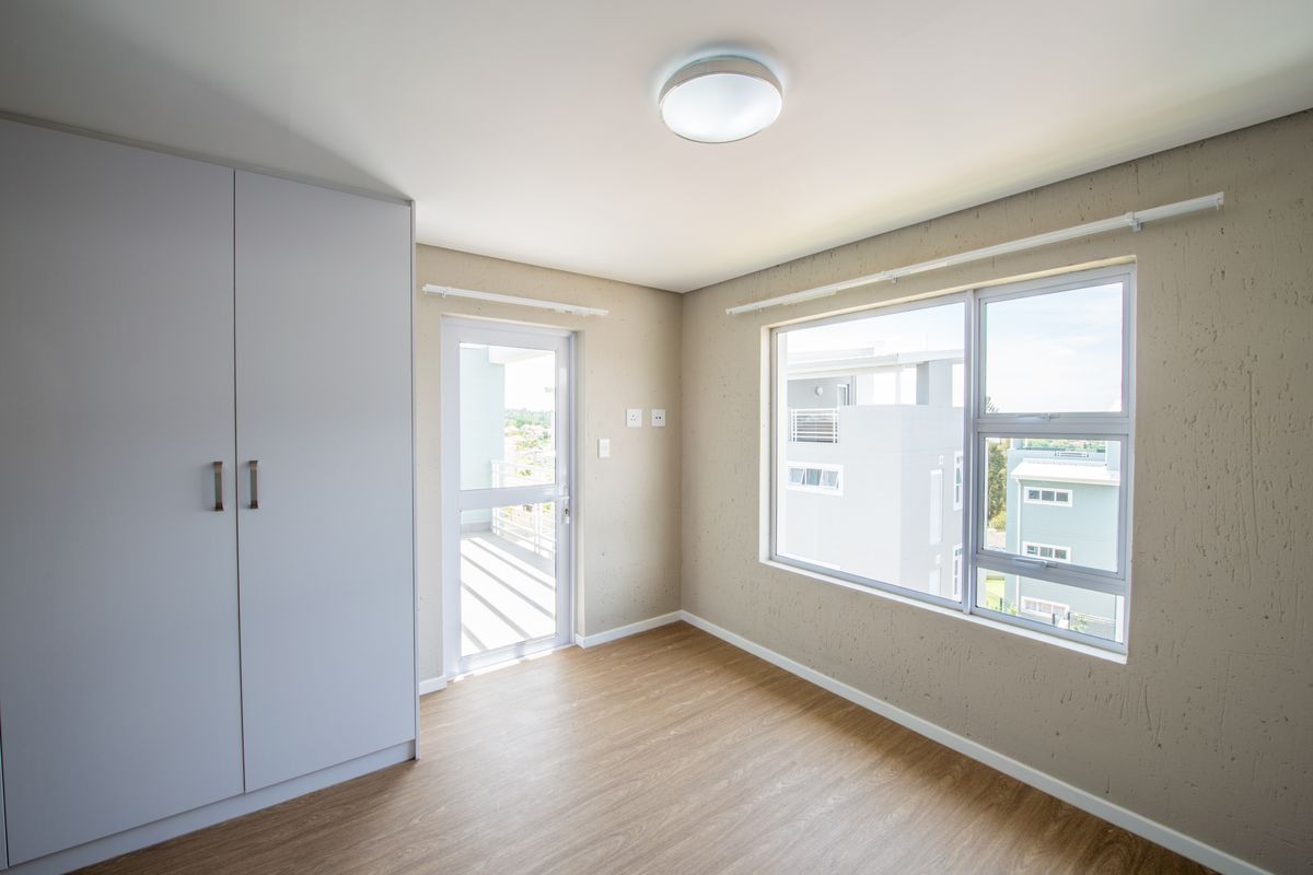 A bright, empty bedroom featuring modern laminate flooring, a built-in wardrobe, and direct access to a private balcony through a glass door.