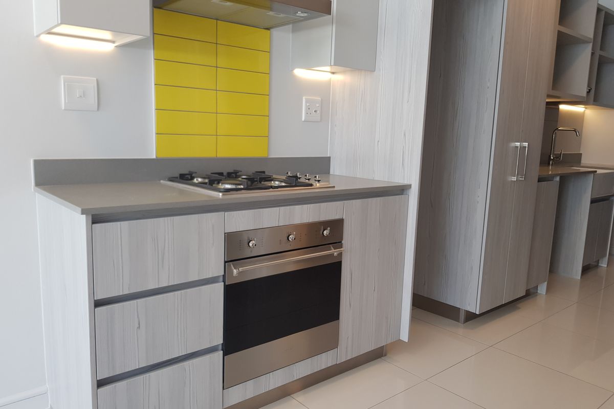 Modern kitchen featuring light wood-grain cabinetry, stone countertops, a gas hob with extractor fan, and a vibrant yellow tiled splashback.