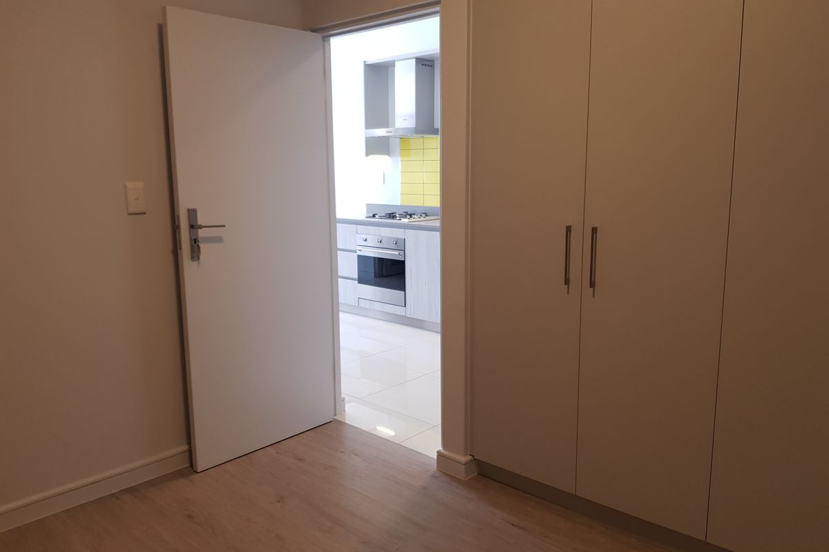 A clean, empty bedroom featuring built-in cupboards and laminate flooring, with a view through the doorway into a modern kitchen with tiled finishes.