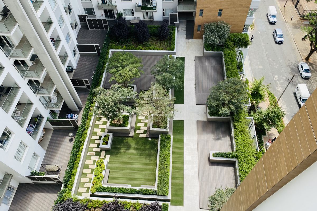 An elevated aerial view of a modern apartment complex featuring a well-maintained landscaped courtyard with timber decking, paved pathways, and manicured garden beds.