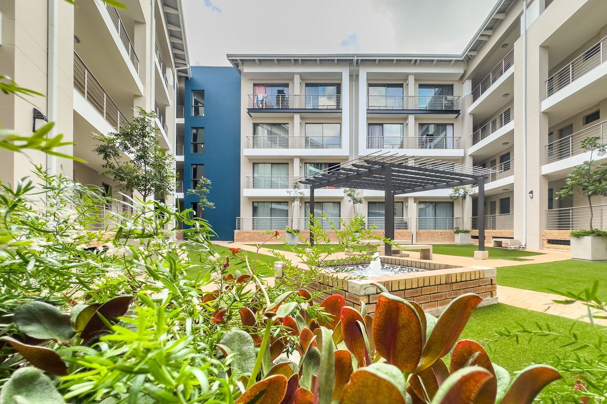 A well-maintained apartment complex courtyard featuring a central water feature, pergola, landscaped garden areas, and private balconies for the residential units.