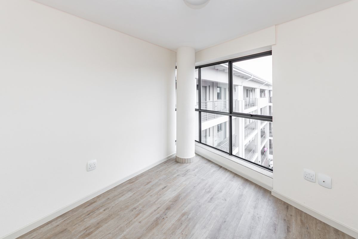 A bright, empty bedroom featuring modern laminate flooring, a structural column, and large windows overlooking the apartment complex.