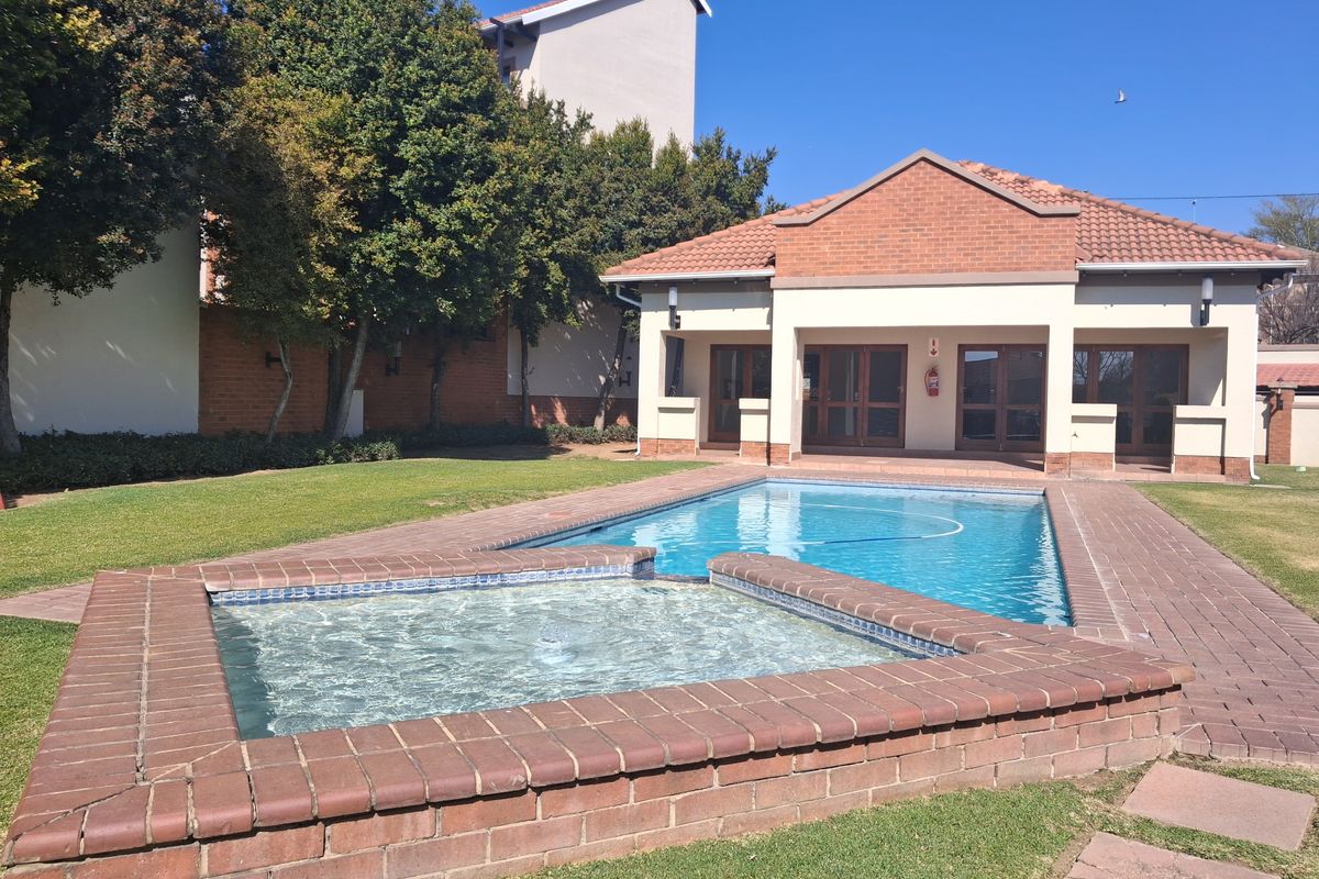 A well-maintained communal pool area featuring a raised spa, brick paving, and an adjacent clubhouse building with a tiled roof.