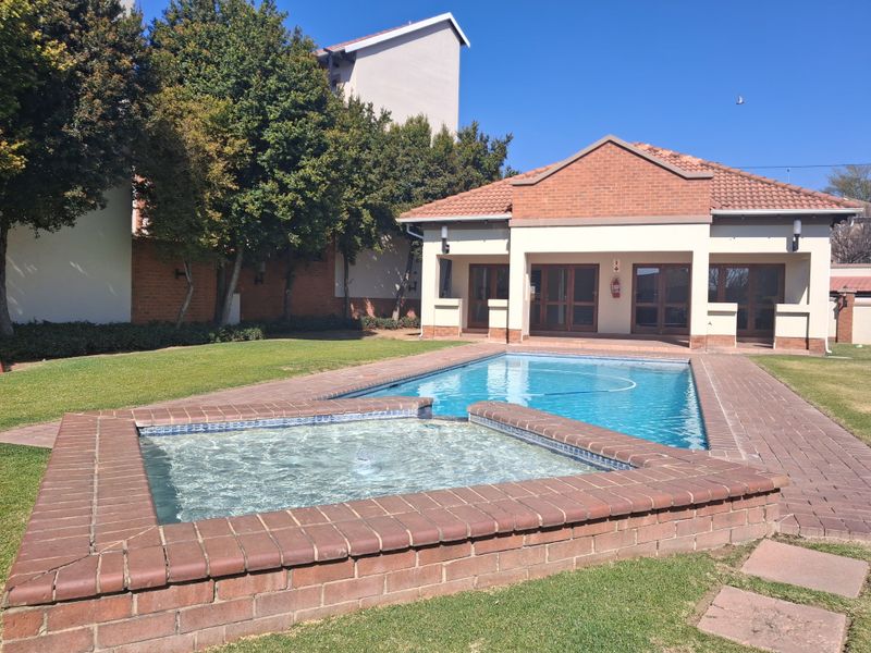 A well-maintained communal pool area featuring a raised spa, brick paving, and an adjacent clubhouse building with a tiled roof.