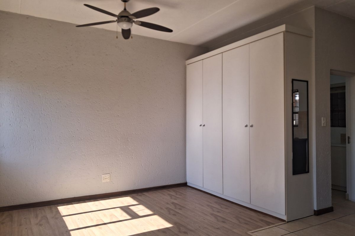 A bright, empty bedroom featuring light laminate flooring, a ceiling fan, and a large white built-in wardrobe with a side mirror.
