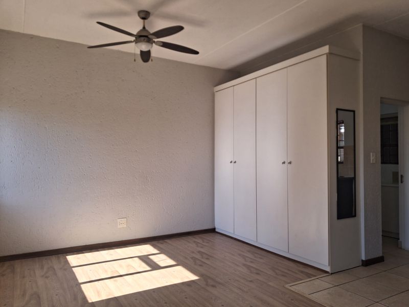 A bright, empty bedroom featuring light laminate flooring, a ceiling fan, and a large white built-in wardrobe with a side mirror.