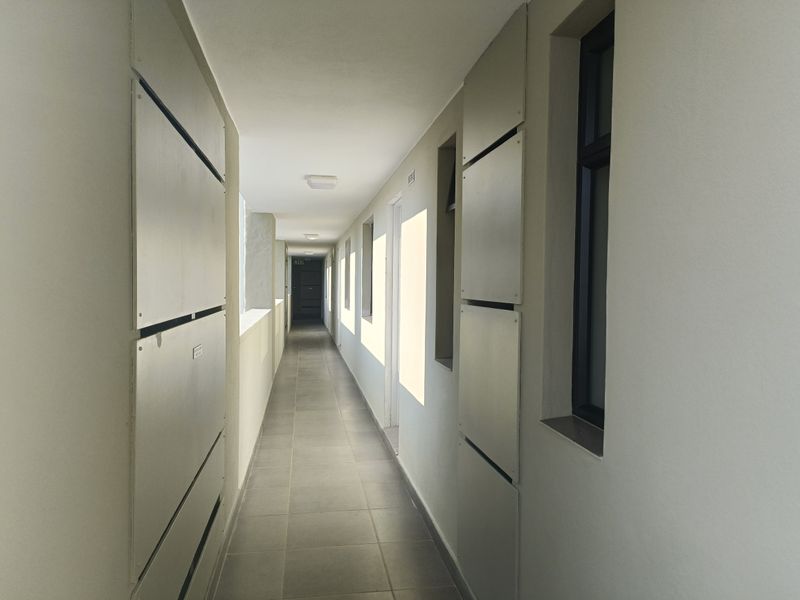 A clean, well-lit apartment hallway featuring neutral tiled flooring, modern service access panels, and multiple window openings providing natural light.