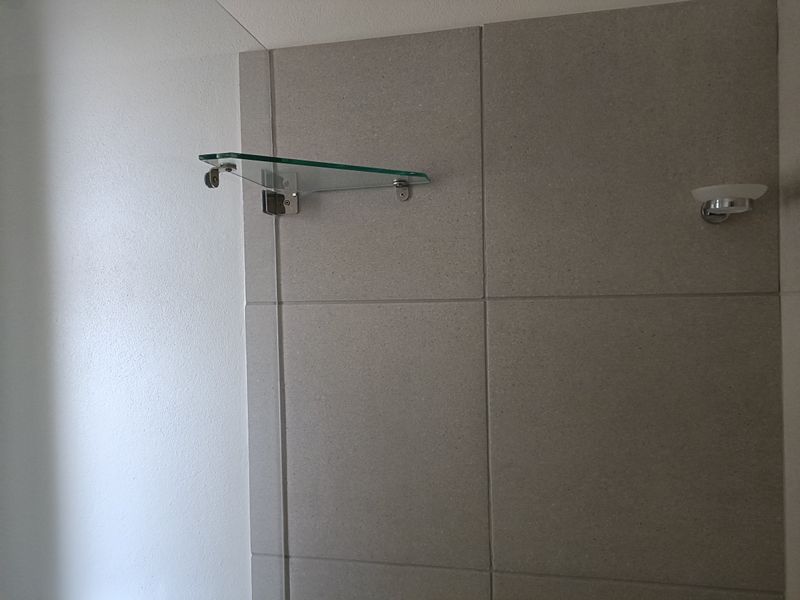 A close-up view of a bathroom wall featuring neutral ceramic tiling, a mounted glass shower shelf, and a small soap dish.
