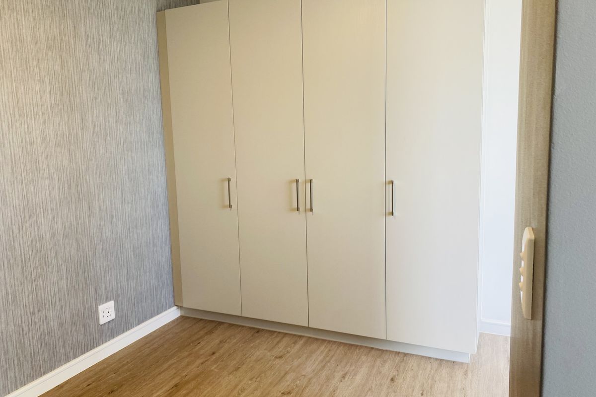 A clean, empty bedroom featuring modern laminate flooring, textured accent wallpaper, and a large, neutral-toned built-in wardrobe with contemporary hardware.