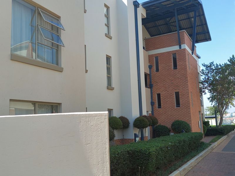 A modern apartment building exterior featuring a combination of brick and plastered finishes, metal roofing, and well-maintained landscaped hedging along a paved walkway.