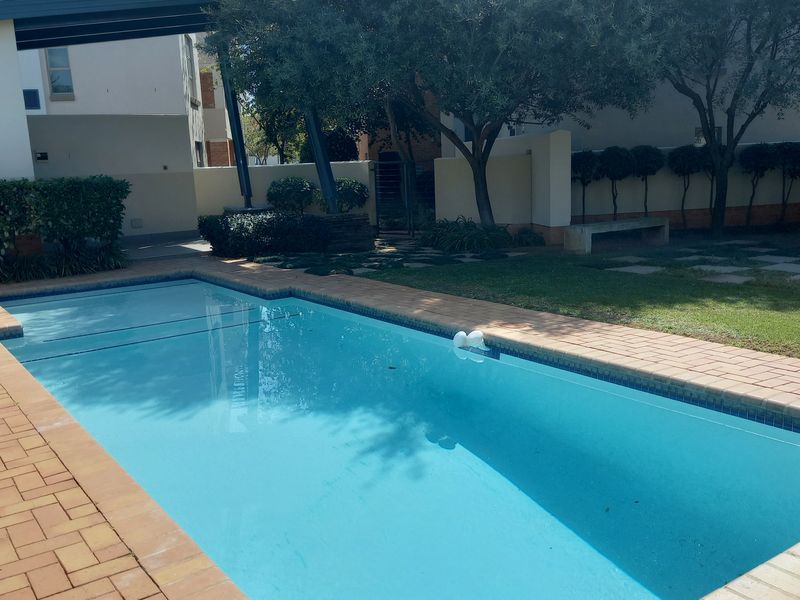 A well-maintained outdoor swimming pool area featuring brick paving, landscaped garden surroundings, and a built-in bench within a residential apartment complex.