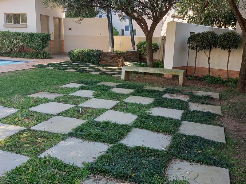 A landscaped garden area featuring a checkerboard paved path, a stone bench, mature trees, and access to a swimming pool.