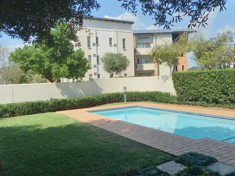 A well-maintained outdoor pool area featuring a swimming pool, brick paving, and landscaped garden borders within a secure apartment complex setting.