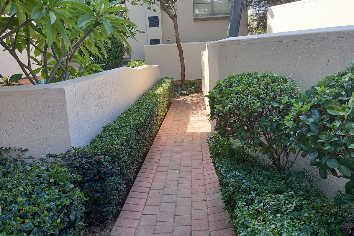 A well-maintained brick paved walkway leading through a landscaped garden area with neat hedges and retaining walls.