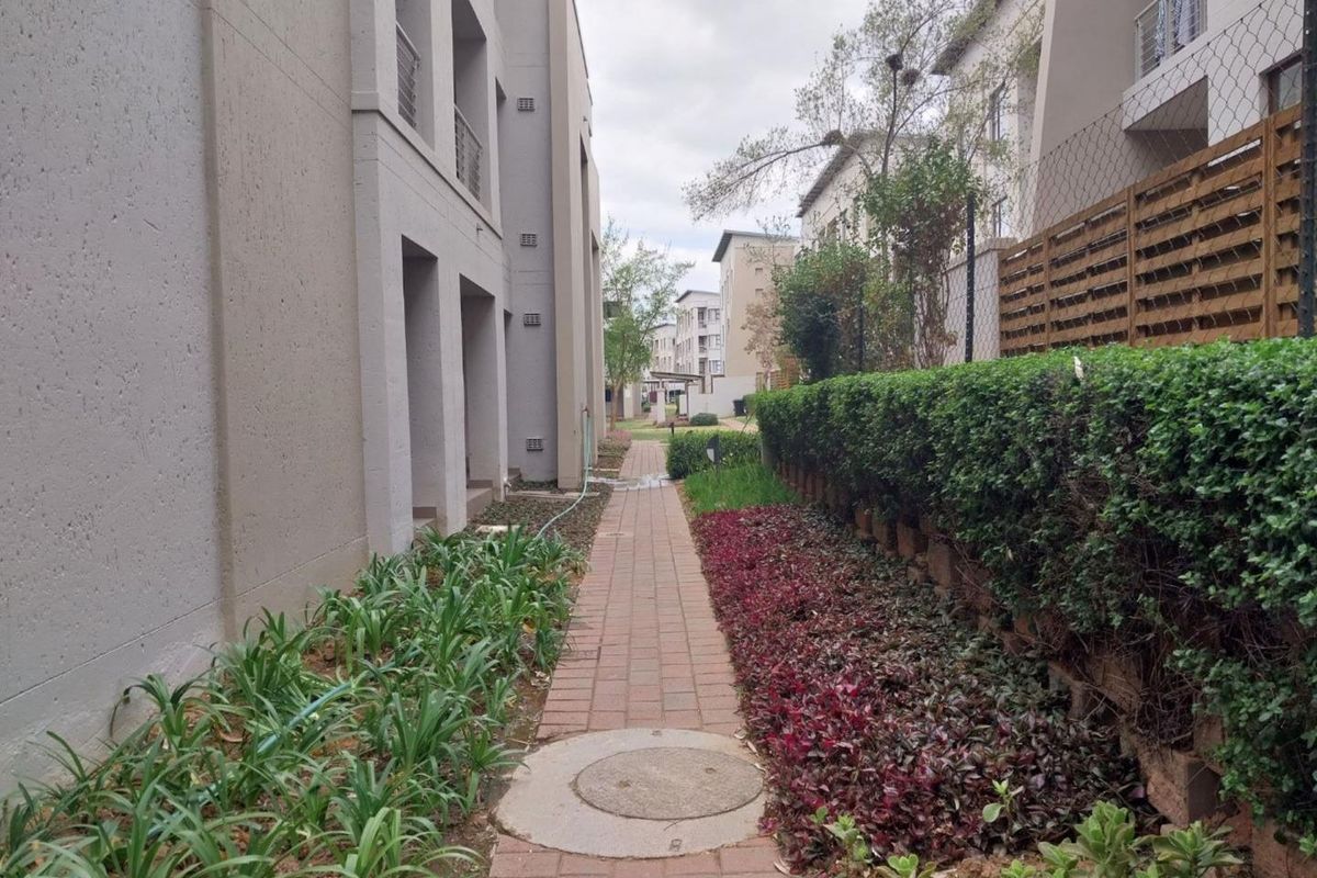 A paved walkway between apartment blocks featuring landscaped garden beds, a retaining wall, and perimeter fencing.