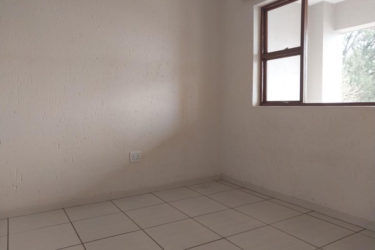 An empty bedroom featuring neutral tiled flooring, textured walls, and a brown aluminium window frame providing natural light.