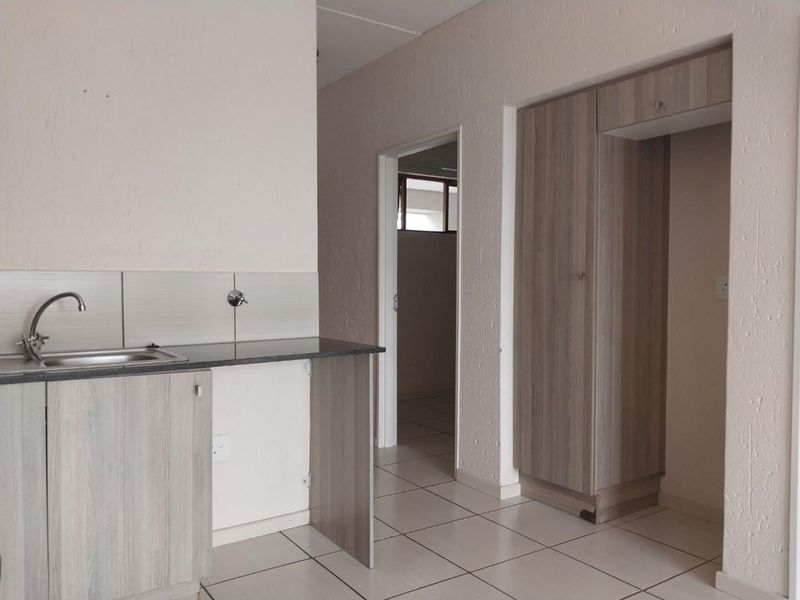 A compact kitchen area featuring light wood-finish cabinetry, a stainless steel sink, and tiled flooring, leading into an adjacent room.