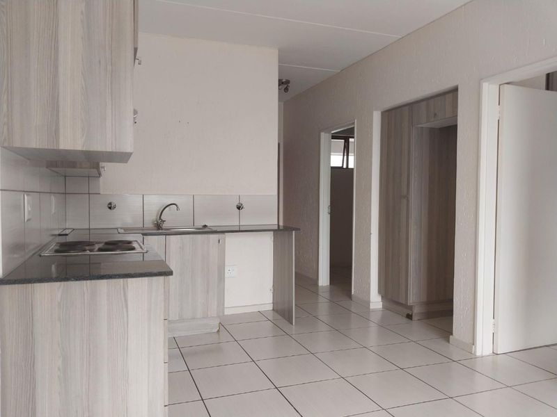 A functional kitchen featuring light wood-look cabinetry, stone countertops, an electric hob, and tiled flooring, leading into a hallway with built-in storage.