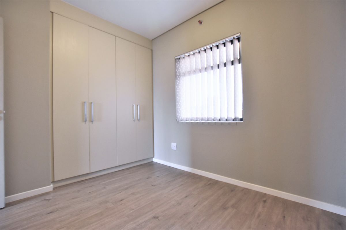 A clean, empty bedroom featuring neutral walls, modern laminate flooring, and ample built-in cupboard storage with vertical blinds on the window.