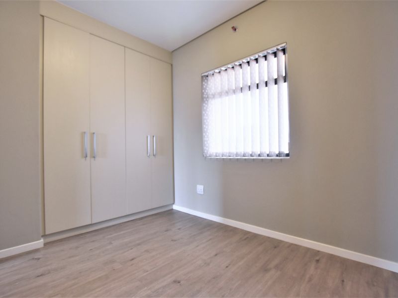 A clean, empty bedroom featuring neutral walls, modern laminate flooring, and ample built-in cupboard storage with vertical blinds on the window.