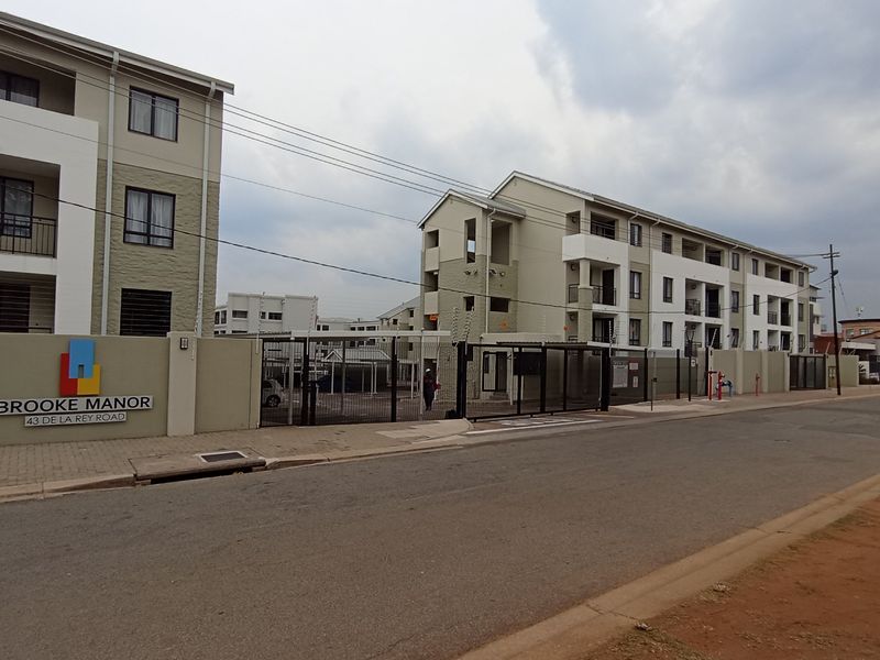 Street view of Brooke Manor apartment complex featuring a secure gated entrance, perimeter wall with electric fencing, and multi-story residential buildings.