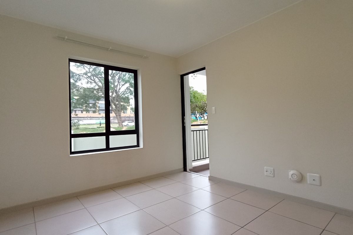 A bright, empty bedroom featuring neutral tiled flooring, clean walls, and direct access to a private balcony through a glass-paneled door.