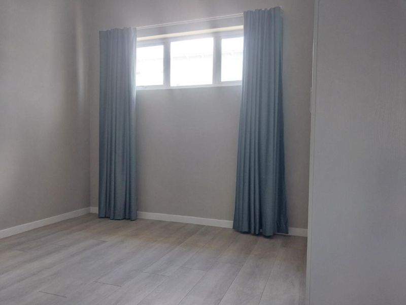 A bright, empty bedroom featuring neutral wall tones, modern laminate flooring, and a window with installed curtain tracks.