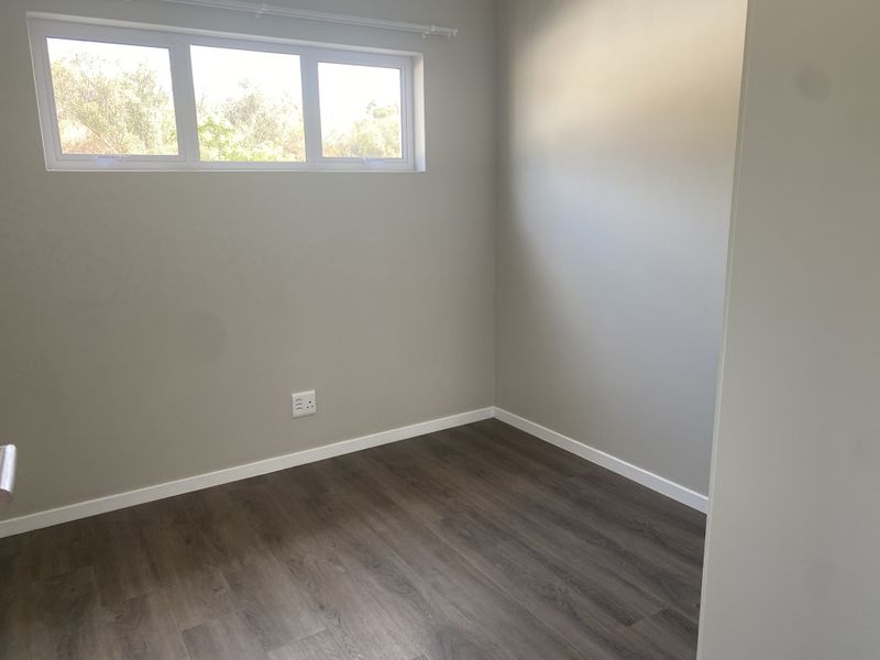 A clean, empty bedroom featuring modern laminate flooring, neutral wall paint, and a high-set window with a curtain rod.