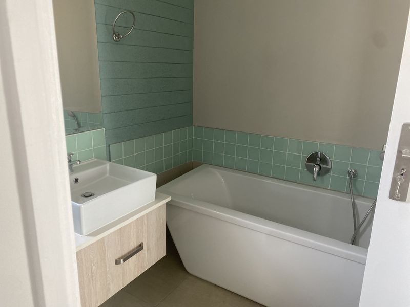 A clean, modern bathroom featuring a white freestanding bathtub, a wall-mounted vanity with a basin, and decorative green tiled wall accents.