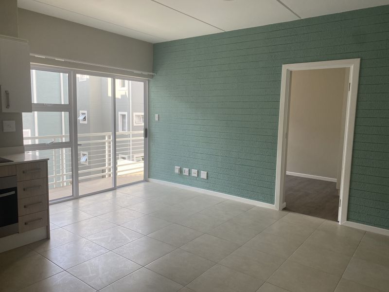 An open-plan living and kitchen area featuring tiled floors, a textured green accent wall, and a sliding door leading to a balcony.